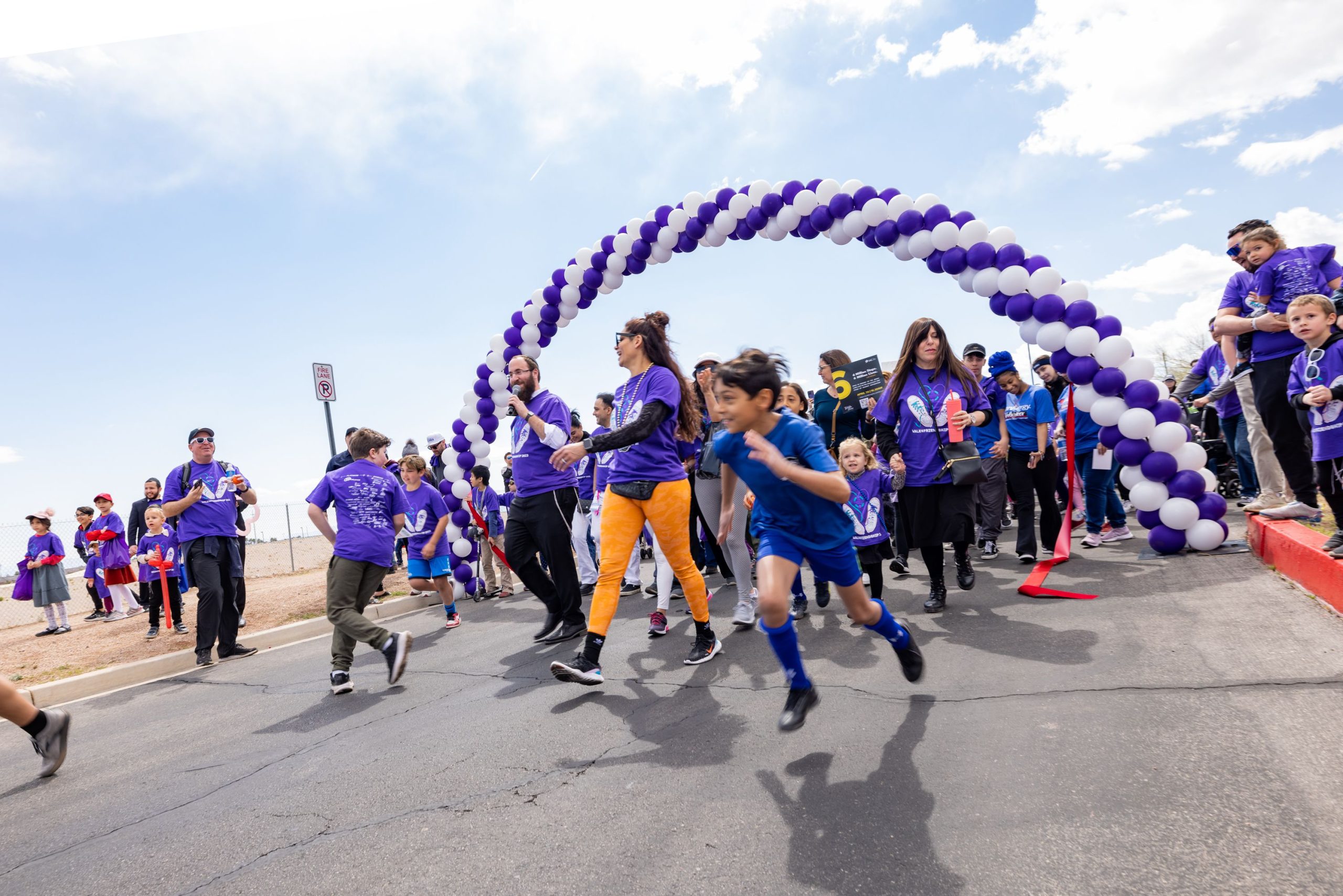 Test Event 1 A crowd of people in purple shirts walk and run under a purple and white balloon arch on a sunny day.