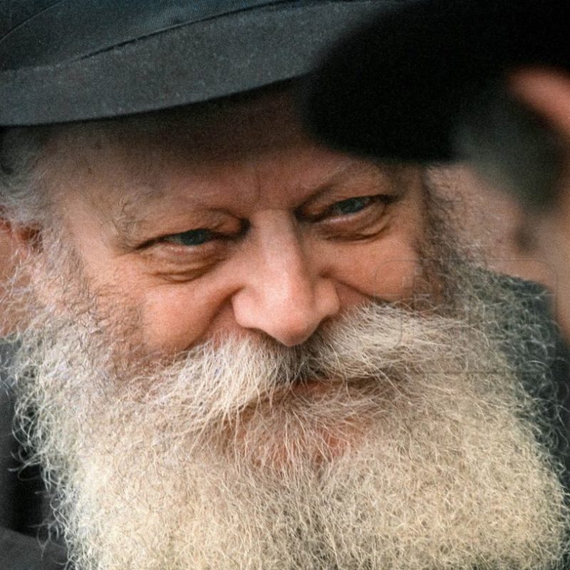 A man with a long, thick white beard and mustache smiles, wearing a dark hat that casts a shadow.